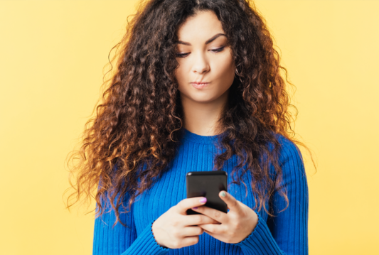 Young woman in a blue shirt looking down at her phone with a pensive expression.