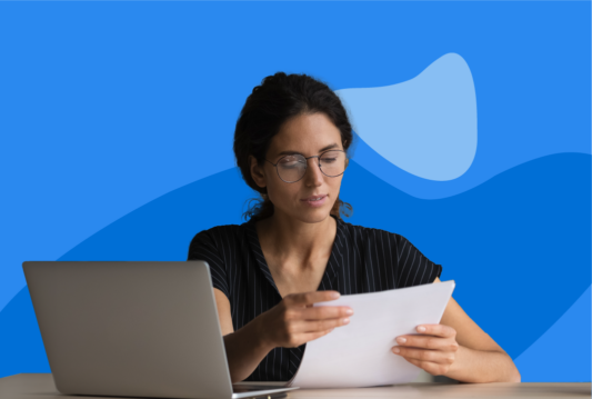 Woman with glasses looking at a piece of paper while sitting in front of a laptop.