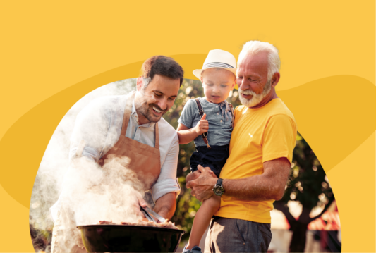 A grandfather holds his grandson while watching the father flip meat on a smoking barbecue on a summer day.