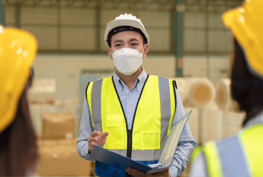 To comply with WHIMS, an employee wearing a high-visibility vest, hard hat, and face mask is addressing a group of employees while holding a binder.
