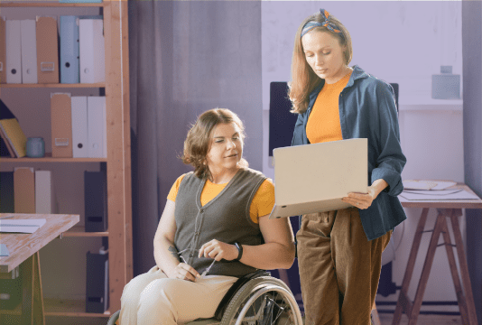 AODA Guidelines: An employee in a wheelchair is talking with another employee who is standing next to them, holding a laptop towards them. They are both looking at the laptop.