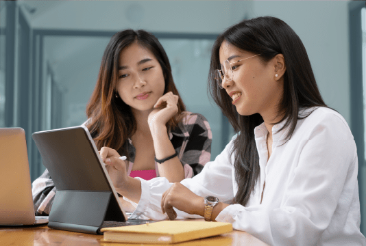 Student employment program: A student employee is sitting next to a work buddy at a desk. They are both smiling and looking at the screen on the desk.