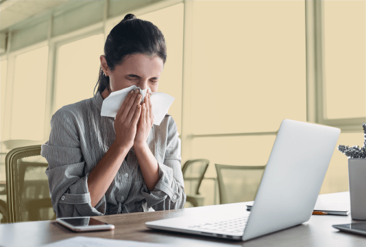 Encouraging sick days: An employee is blowing their nose while working at their desk, looking at their laptop