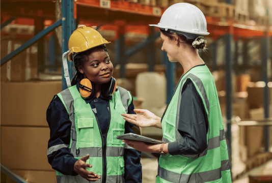 Workplace Wellness: A worker has a tablet and is talking to another employee in a warehouse. They are both wearing hard hats and discussing safety controls.